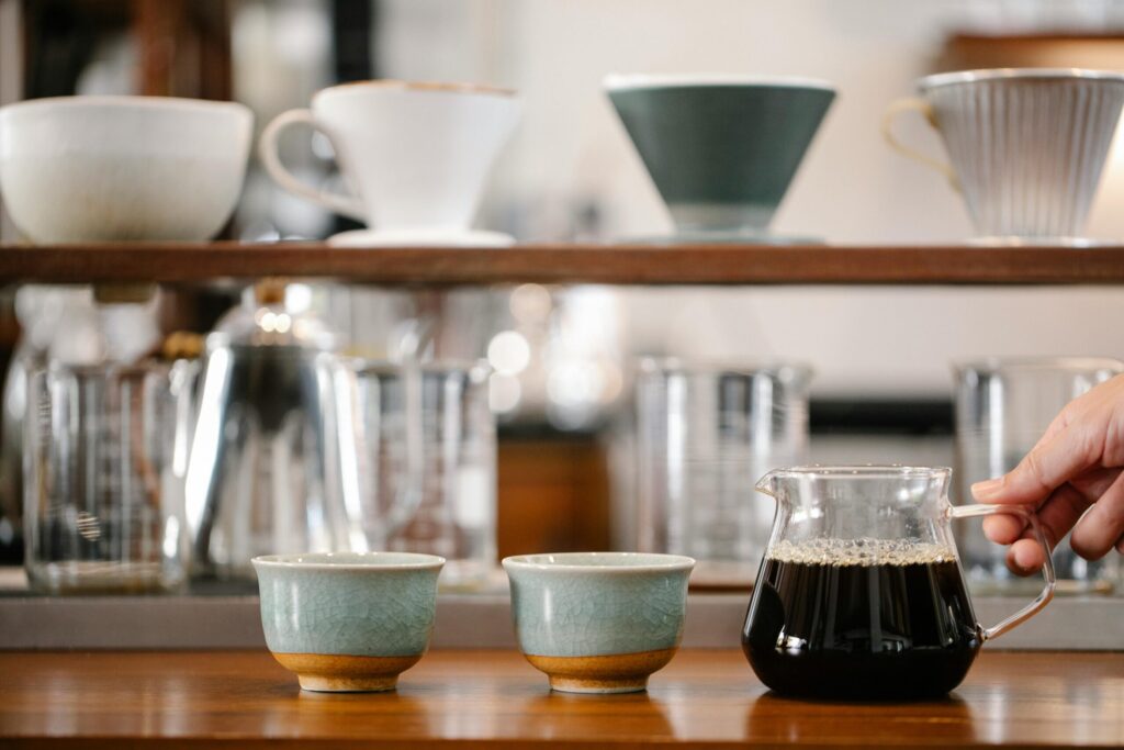 Crop anonymous barista putting glass pot with coffee on table with cups near beakers and drippers on blurred background in modern cafe